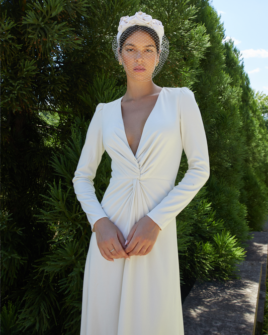 First image of Misha Dress, a white wedding maxi dress with long sleeves, high slit in middle, twist detail in the front, plunging v-neckline, lining, and back zipper